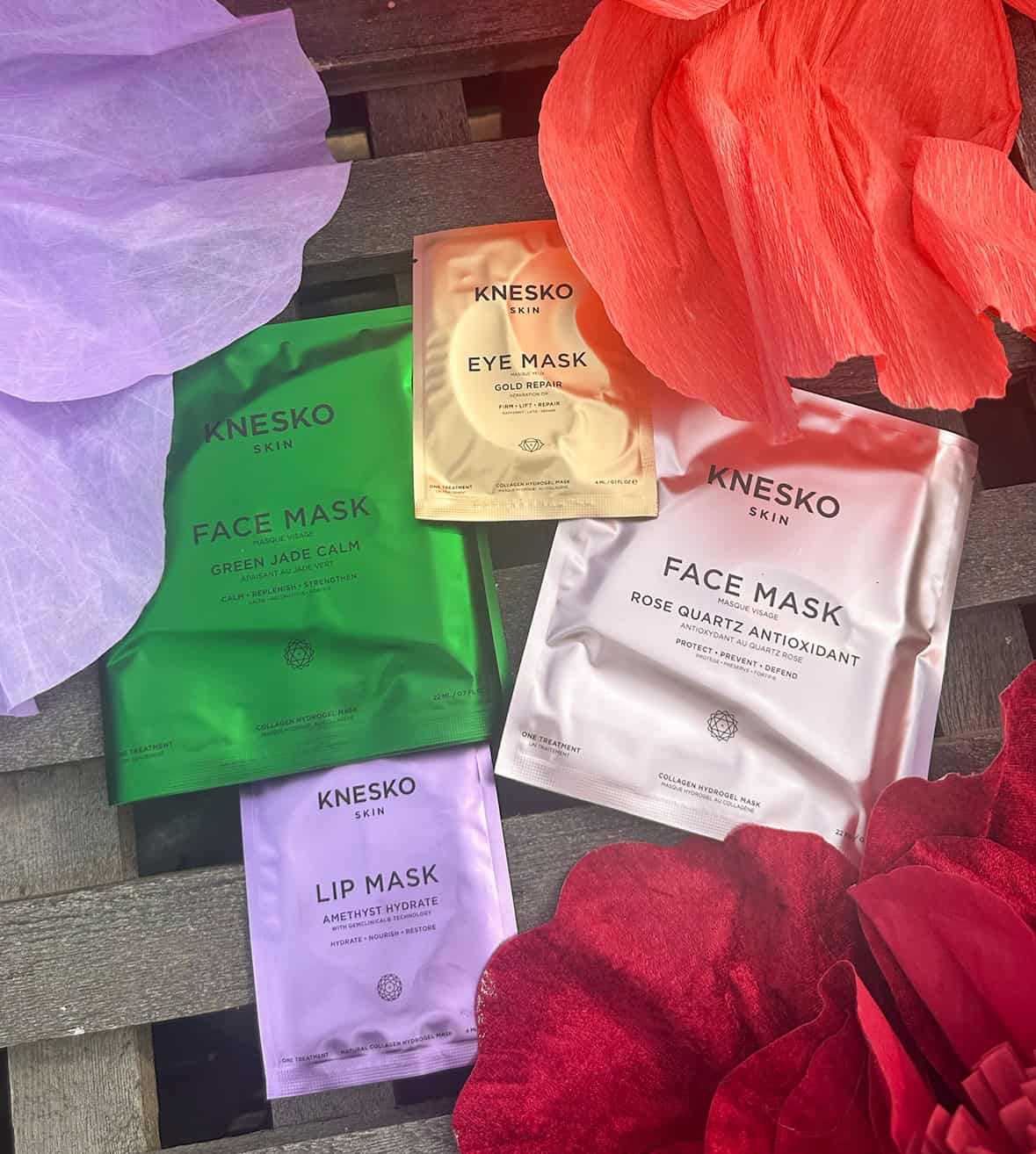 Assorted Knesko skin face masks and eye mask on wooden surface with flower petals.